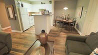 This single-floor space showcases an open-concept layout that effortlessly connects the living, dining, and kitchen areas into one cohesive and functional environment. Rich, warm-toned hardwood-style flooring runs throughout, lending continuity and a welcoming ambiance to the entire space.

The kitchen is outfitted with crisp white cabinetry that extends to the upper walls, offering generous storage. A white appliance suite, including a refrigerator and range, complements the clean aesthetic. A peninsula-style counter serves as both a prep space and a natural visual divider between the kitchen and the adjoining areas, with room for casual seating or additional workspace.

Adjacent to the kitchen, the dining area is anchored by a wood-toned rectangular table paired with dark metal-framed chairs, creating an inviting spot for meals. A pendant light hangs above, providing focused illumination over the dining space.

The living area flows naturally from the dining zone, featuring a comfortable seating arrangement centered around a glass-topped coffee table adorned with decorative accents. The walls are finished in a soft, neutral green-gray tone that adds warmth without overwhelming the space.

Overall, the layout is practical and thoughtfully designed, offering an easy, connected flow between everyday living spaces, ideal for both relaxed daily living and casual entertaining.