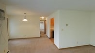 This single-floor residence offers a straightforward and functional layout designed for comfortable everyday living. The main living and dining area is finished with neutral wall-to-wall carpeting, creating a warm and cohesive feel throughout the space. A decorative pendant light fixture hangs in the dining area, adding a classic touch to the open room. The interior is finished with clean white walls and light-toned baseboards that contribute to a bright, airy ambiance. A partial wall divides the main living area from the kitchen, which features warm wood cabinetry and what appears to be dark appliances, providing a practical and organized cooking space with its own distinct flooring. A hallway extends toward the back of the residence, connecting the main living spaces to additional rooms, with a doorway visible at the far end suggesting a bedroom or utility area. The layout flows naturally from the living and dining zone through to the kitchen and private spaces, making the overall floor plan easy to navigate. Ceiling-mounted lighting in the hallway ensures the passage remains well-lit. The neutral color palette and consistent finishes throughout give the space a clean, move-in-ready appearance.