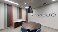 This single-floor conference room is thoughtfully designed to support focused collaboration and professional meetings. At the center of the room sits a circular walnut-toned conference table surrounded by blue upholstered rolling chairs, creating an inclusive, equal-footing meeting environment. A bold statement pendant light in matte black hangs directly above the table, providing both functional illumination and a design focal point.

One wall features a striking vertical striped accent design in blue, orange, and yellow tones, adding a vibrant and dynamic character to the otherwise neutral, white-walled interior. Adjacent to this accent wall is a built-in credenza with dark cabinetry and a light granite countertop, offering useful surface and storage space.

A distinctive row of analog wall clocks lines one wall, displaying multiple time zones — a practical and visually engaging feature that signals the room's use for international or multi-regional coordination. The room also includes a large interior window, allowing visibility into an adjacent space, along with a glass-paneled door that enhances transparency and openness. Neutral carpet tiles cover the floor throughout, and recessed and linear ceiling lighting ensures consistent, well-distributed illumination across the entire room.