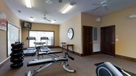 This fitness room offers a functional and well-organized space for exercise, finished with warm beige walls, dark wood baseboards, and carpet flooring that lend it a residential yet purposeful character. Two ceiling fans circulate air throughout the room, contributing to a comfortable workout environment. Natural light enters through a large window fitted with horizontal blinds, brightening the space alongside overhead fluorescent lighting. The room is equipped with multiple treadmills positioned near the window, along with a weight bench and additional exercise equipment, accommodating both cardio and strength training needs. A round wall mirror is mounted on one wall, a practical feature for monitoring form during workouts. A small console table holds basic amenities, and a wall-mounted information board is also present. Two dark wood panel doors provide access to adjoining areas, maintaining a cohesive aesthetic throughout. The room's layout allows for comfortable movement between equipment, making it a practical and inviting space for daily fitness use.