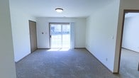 This single-floor space offers a straightforward and functional layout designed for comfortable everyday living. The main living area is defined by plush wall-to-wall carpeting in a neutral tone, providing a soft and inviting underfoot experience. Clean white walls keep the interior feeling bright and open, while flush-mount ceiling lighting ensures consistent illumination throughout the room. A standout feature of the living area is the sliding glass door, framed in warm wood tones and dressed with sheer white vertical blinds. This element serves a dual purpose — flooding the space with abundant natural light and providing direct access to the outdoors, where a green lawn is visible beyond. The entry door, finished in a classic wood tone, is conveniently positioned adjacent to the main living area, contributing to an efficient and accessible floor plan. White baseboard trim runs along the perimeter of the room, adding a clean finishing detail that complements the overall neutral color palette. The space is characterized by its simplicity, practicality, and the seamless connection between the interior living environment and the natural outdoor setting, making it a welcoming and functional place to call home.