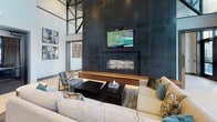 The REVA Main St Rahway Resident Lounge in Rahway, NJ, is a thoughtfully designed communal space that blends modern aesthetics with functional amenities. The lounge's most striking architectural feature is its soaring double-height ceiling, accented by bold steel-framed clerestory windows that flood the interior with natural light. A dramatic floor-to-ceiling dark tile accent wall serves as the focal point of the main lounge area, anchoring a linear gas fireplace with a live-edge wood hearth bench and a mounted flat-screen television above. Comfortable sectional seating and curated artwork create an inviting atmosphere for relaxation.

A dedicated dining and co-working area features a slatted wood partition wall with integrated lighting, built-in banquette seating, and long communal tables suited for both casual dining and productive work sessions. Globe pendant fixtures and abstract wall murals add artistic character throughout.

The kitchen bar area is outfitted with dark cabinetry, hexagonal tile flooring, bar-height seating, and a coffee station. Large black-framed glass doors open directly to an outdoor courtyard, seamlessly connecting the interior to exterior amenity spaces. Light wood flooring, sculptural pendant lighting, and carefully selected décor complete the lounge's polished, contemporary ambiance.