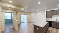 This single-floor space showcases a clean, modern design with an open-concept layout that seamlessly connects the kitchen and living areas. The kitchen features a prominent island topped with speckled granite countertops and dark espresso cabinetry with sleek stainless steel hardware, providing both style and functional prep space. The living area is expansive and airy, bathed in natural light streaming through a full-width sliding glass door fitted with vertical blinds, which opens to an outdoor space. The flooring throughout is a warm hardwood-style laminate that adds continuity and a sense of warmth to the open floor plan. Recessed lighting is thoughtfully distributed across the white ceiling, ensuring bright, even illumination throughout the space. The walls are finished in a neutral off-white tone, creating a versatile backdrop suited to a variety of interior styles. A half-wall partition near the entryway subtly defines the transition between the entry corridor and the main living space, adding architectural interest without sacrificing the open feel. Overall, the space presents a bright, well-appointed, and move-in-ready environment with a contemporary aesthetic.