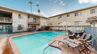 This multi-unit residential complex is designed around a vibrant, open-air courtyard that serves as the heart of the community. At the center of the courtyard sits a generously sized swimming pool with clear, blue water, surrounded by a tiled sun deck furnished with chaise lounges, patio chairs, and shade umbrellas — ideal for outdoor relaxation and recreation.

The complex features a stucco exterior finished in warm beige tones, with multi-level balconies accessible via interior staircases and walkways. Ground-floor units open directly onto covered patios, while upper-level units enjoy elevated balconies with decorative iron railings, offering views of the courtyard below. Sliding glass doors connect interior living spaces to the outdoor areas.

Security is a key feature of the property, with blue metal-gated entry points securing the pool area and courtyard perimeter. Manicured shrubs and potted plants are placed throughout, adding greenery and a welcoming ambiance. Tall palm trees rise above the roofline, reinforcing a distinctly Southern California aesthetic and providing a sense of lush, resort-like character.

The open-sky courtyard layout allows for abundant natural light throughout the day, creating a bright and airy atmosphere. The overall design emphasizes communal outdoor living, with the pool and shared spaces positioned to foster a connected, community-oriented environment.