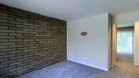 This multi-floor space makes a distinct visual impression through its combination of rustic and understated design elements. The most prominent architectural feature is a full exposed brick accent wall, rendered in earthy, dark-toned brick laid in a classic running bond pattern. This wall serves as a strong focal point, introducing texture and character that contrasts with the surrounding smooth, white-painted walls. The flooring throughout is soft, wall-to-wall carpet in a neutral grey-blue tone, contributing to a comfortable and quiet atmosphere underfoot. The ceiling features a textured popcorn finish, a detail consistent across the visible areas of the space. The overall color palette is restrained and neutral, allowing the brick wall to command attention while keeping the remainder of the space open and adaptable. The layout across three floors provides versatility in how the space can be organized and utilized. Clean white walls offer a blank canvas for personalization, and the combination of the tactile brick surface, plush carpeting, and textured ceiling gives the space a layered, cozy quality that balances both warmth and functionality.