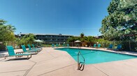This outdoor recreational space centers around a large, freeform swimming pool with clear turquoise water, designed to accommodate both leisurely swimming and relaxation. The pool features accessible entry steps with stainless steel handrails and is clearly depth-marked for safety. Surrounding the pool is a broad, open concrete sun deck that provides ample room for lounging and circulation.

The deck is furnished with multiple teal-colored chaise lounges and accompanying side tables, arranged to take full advantage of the abundant natural sunlight. Several patio umbrella tables offer shaded seating areas for those seeking relief from the sun, making the space suitable for both sunbathing and casual outdoor dining.

The entire pool area is enclosed by a sturdy metal perimeter fence with a gated entry, ensuring both security and privacy. Mature trees — including tall pines and broad deciduous varieties — line the boundaries of the space, providing natural shade, visual screening, and a lush, park-like ambiance. Well-maintained shrubs and greenery further enhance the landscaping throughout.

A multi-story residential building is visible beyond the fencing, indicating this pool serves as a shared amenity. The overall setting is clean, well-maintained, and designed for maximum outdoor enjoyment under open skies.
