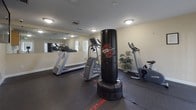 This dedicated fitness room is designed to support a comprehensive workout experience within a compact, single-floor layout. The space features durable dark rubber flooring throughout, providing a safe and functional surface for a wide range of exercises. A large wall-mounted mirror spans one side of the room, visually expanding the space and allowing users to monitor their form during workouts.

The room is outfitted with a variety of equipment, including a treadmill, a stationary recumbent bike, a multi-station strength training machine, and a freestanding Wave Master punching bag, catering to both cardio and strength training needs. A flat-screen television is mounted on the wall, offering entertainment during workouts. Framed exercise guides are also displayed on the wall, providing instructional references for users.

Natural light filters in through multiple windows fitted with horizontal blinds, complementing the overhead flush-mount lighting to keep the space bright and welcoming. The neutral beige walls contribute to a clean, focused atmosphere. The room also includes safety essentials such as a fire extinguisher and an illuminated emergency exit sign. Two exterior doors provide convenient access points, making this fitness room a practical and well-appointed amenity.