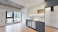 Located at 250 Rue Young in Montréal's dynamic Griffin town neighbourhood, this efficiently designed unit offers a thoughtful layout that maximizes every square metre of its single-floor plan. Light oak hardwood flooring flows throughout, complemented by crisp white walls and a striking exposed concrete ceiling that lends an industrial-modern character to the living area. A large floor-to-ceiling window floods the main space with natural light while framing views of the surrounding cityscape.

The kitchen is a standout feature, outfitted with two-tone cabinetry — matte slate-blue lower cabinets paired with cream upper cabinets — and under-cabinet LED lighting. Vertical subway tile forms a clean backsplash behind the integrated cooktop, and stainless steel appliances including an oven, dishwasher, and full-size refrigerator provide full functionality. A wall-mounted air conditioning unit services the main living area for year-round comfort.

In-unit stacked washer and dryer are neatly tucked behind a sliding door, keeping the living space uncluttered. The bathroom is finished to a high standard, featuring large-format floor tiles, a frameless sliding glass shower enclosure with full-height tile surround, a wall-mounted medicine cabinet with mirror, and a warm wood-toned vanity with an undermount sink.