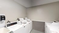 This shared laundry facility is a practical, single-floor space designed to accommodate multiple users efficiently. The room is finished with neutral gray walls and durable light-toned flooring, creating a clean and functional environment throughout.

Multiple commercial-grade top-load washers and dryers are arranged along the walls, providing ample laundry capacity. A spacious laminate folding counter runs along one wall, offering a convenient surface for sorting and folding laundry. A deep utility sink is positioned nearby, ideal for hand-washing or pre-treating garments. Wall-mounted paper towel and soap dispensers add to the room's practicality and hygiene.

The space is equipped with key safety features, including a prominently mounted fire extinguisher and a fire alarm pull station, ensuring occupant safety. A glazed exterior door allows natural light to enter the room and provides direct outdoor access. Overhead lighting keeps the space well-lit at all times.

A waste bin is conveniently placed for easy disposal, and the coin-operated machines indicate this is a shared facility. The overall layout is thoughtfully organized to allow comfortable movement between machines and workspaces, making it a fully functional and well-maintained communal laundry room.