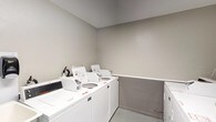 This shared laundry facility is a practical, single-floor space designed to accommodate multiple users efficiently. The room is finished with neutral gray walls and durable light-toned flooring, creating a clean and functional environment throughout.

Multiple commercial-grade top-load washers and dryers are arranged along the walls, providing ample laundry capacity. A spacious laminate folding counter runs along one wall, offering a convenient surface for sorting and folding laundry. A deep utility sink is positioned nearby, ideal for hand-washing or pre-treating garments. Wall-mounted paper towel and soap dispensers add to the room's practicality and hygiene.

The space is equipped with key safety features, including a prominently mounted fire extinguisher and a fire alarm pull station, ensuring occupant safety. A glazed exterior door allows natural light to enter the room and provides direct outdoor access. Overhead lighting keeps the space well-lit at all times.

A waste bin is conveniently placed for easy disposal, and the coin-operated machines indicate this is a shared facility. The overall layout is thoughtfully organized to allow comfortable movement between machines and workspaces, making it a fully functional and well-maintained communal laundry room.