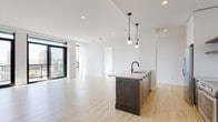 This modern single-floor residence offers a thoughtfully designed open-concept layout that seamlessly integrates the living, dining, and kitchen areas into one cohesive, light-filled space. Light wood-tone flooring runs throughout, complementing the crisp white walls and recessed lighting that give the unit a clean, contemporary feel.

The kitchen is a standout feature, anchored by a substantial dark-cabinetry island with a light countertop, an undermount sink, and a matte black faucet. Pendant lights hang above the island, adding a refined touch. The perimeter kitchen wall is equipped with two-tone cabinetry — white upper cabinets paired with dark lower units — a white tile backsplash, and stainless steel appliances including a range and over-the-range microwave.

A dedicated laundry closet houses a stacked washer and dryer unit alongside a water heater, providing practical in-unit utility. The bedroom offers a quiet, neutral retreat with the same warm flooring and ample wall space. The bathroom features large-format stone-look floor tiles, white subway tile surrounding a soaking tub and shower, and sleek matte black fixtures throughout.

Floor-to-ceiling sliding glass doors open to a private balcony, bringing in abundant natural light and offering expansive urban skyline views, making this a standout feature of the living area.