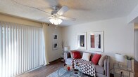 This single-floor space offers a comfortable and well-appointed living environment designed with both style and functionality in mind. The living room serves as the heart of the space, showcasing warm neutral walls that complement the rich wood-look flooring running throughout. A multi-blade ceiling fan with an integrated light fixture provides both ambient lighting and year-round climate comfort. Large vertical-blind-covered sliding glass doors flood the room with natural light while offering privacy and access to the outdoors. The living area is thoughtfully arranged with a plush sofa accented by red and patterned throw pillows, centered around a sleek glass coffee table that adds a modern touch. Framed wall art adds personality and visual interest to the space. The textured ceiling adds a classic architectural detail consistent throughout. Overall, the space exudes a warm, inviting ambiance with a harmonious blend of contemporary furnishings and traditional design elements, making it a comfortable retreat suited for everyday living and relaxed entertaining.