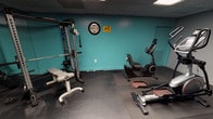 This fitness room is a purpose-built exercise space designed to accommodate a variety of workout styles. The room features bold teal accent walls contrasted against darker gray walls, creating an energetic and motivating atmosphere. A drop-panel ceiling with recessed flat lighting provides consistent, even illumination throughout the space.

The room is outfitted with a solid selection of cardio equipment, including a treadmill, an elliptical trainer, and a recumbent stationary bike, each positioned on protective rubber floor mats. For strength training, a multi-function cable and weight machine is available alongside a workout bench, making it suitable for a full-body exercise regimen. A stability ball rounds out the available fitness accessories.

A wall-mounted flat-screen television is positioned to be visible from the cardio machines, offering entertainment during workouts. A wall clock and a security camera notice are also present, adding to the practical and safety-conscious setup of the room. The dark rubberized flooring throughout provides excellent traction and durability. Overall, this fitness room offers a functional and well-organized environment for users seeking a convenient on-site workout facility.