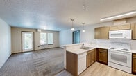 Located at 6930 W Silverado St in Rathdrum, Idaho, this single-floor, one-bedroom, one-bathroom unit offers a practical and comfortable layout. The open-concept kitchen and living area form the heart of the space, with the kitchen featuring warm wood cabinetry, a peninsula countertop with pendant lighting overhead, light hardwood-style flooring, and white appliances including a refrigerator, electric range, and built-in microwave. The peninsula provides a natural divide between the kitchen and the carpeted living room, which receives natural light through windows and a glass door that opens to a small balcony with outdoor views of the surrounding landscape. The bedroom is a well-proportioned retreat, featuring plush carpet, a sliding-door closet, a wall-mounted heating unit, and a window that frames views of nearby mountains and greenery. The full bathroom is cleanly appointed with a dark wood vanity, a marble-toned tile floor, a wide mirror, and a full shower-tub combination. Throughout the unit, neutral tones, textured ceilings, and consistent trim details create a cohesive and understated aesthetic well-suited to everyday living.