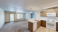 Located at 6930 W Silverado St in Rathdrum, Idaho, this single-floor, one-bedroom, one-bathroom unit offers a practical and comfortable layout. The open-concept kitchen and living area form the heart of the space, with the kitchen featuring warm wood cabinetry, a peninsula countertop with pendant lighting overhead, light hardwood-style flooring, and white appliances including a refrigerator, electric range, and built-in microwave. The peninsula provides a natural divide between the kitchen and the carpeted living room, which receives natural light through windows and a glass door that opens to a small balcony with outdoor views of the surrounding landscape. The bedroom is a well-proportioned retreat, featuring plush carpet, a sliding-door closet, a wall-mounted heating unit, and a window that frames views of nearby mountains and greenery. The full bathroom is cleanly appointed with a dark wood vanity, a marble-toned tile floor, a wide mirror, and a full shower-tub combination. Throughout the unit, neutral tones, textured ceilings, and consistent trim details create a cohesive and understated aesthetic well-suited to everyday living.