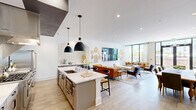 Located at 120 Whitby Lane in Birmingham, Alabama, this single-floor space seamlessly blends a chef's kitchen with an expansive living area, creating a cohesive and sophisticated environment. The living room is anchored by dramatic floor-to-ceiling windows that flood the space with natural light and offer open views of an outdoor patio and surrounding landscape. A tufted cognac leather sofa, a daybed, and a sleek concrete coffee table furnish the seating area, while decorative wooden cutting boards adorn the crisp white walls alongside a large mounted display screen. Striking natural stone accent walls with arched built-in shelving add warmth and architectural character, connecting the living area to additional lounge and game spaces visible in the open layout. The kitchen is outfitted with professional-grade stainless steel appliances, including a commercial-style range with an industrial hood and a French door refrigerator. Light wood cabinetry with dark hardware lines both sides of the galley-style kitchen, topped with white quartz countertops. A central island with an undermount sink and pendant lighting provides additional prep space, while handmade-style tile backsplash and light hardwood floors tie the entire space together with a refined yet inviting aesthetic.