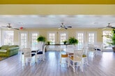 Located at 8181 Colony Dr in Houston, TX, this single-floor community clubhouse offers a generously sized, open-concept gathering space designed for socializing and relaxation. The interior is characterized by warm yellow accent walls, white wainscoting, and light wood-look flooring that runs throughout, creating a cohesive and cheerful atmosphere.

The main gathering area features multiple round white dining tables with white lattice-back chairs, ceiling fans, and an abundance of natural light streaming through numerous French doors and windows. These doors open to views of a pool area, seamlessly blending indoor and outdoor living. Lush potted ferns and tropical plants placed along a long wooden console table and throughout the space enhance the resort-like ambiance.

A dedicated lounge area showcases a bold tropical leaf wall mural as a dramatic backdrop, complemented by green leather sofas, a colorful swirl-patterned area rug, and vibrant yellow accent chairs — creating an inviting, energetic retreat.

The kitchenette is equipped with granite countertops, white cabinetry, a refrigerator, microwave, and sink, with bar-height seating at the peninsula. A vaulted ceiling with linear skylights adds architectural interest and draws in additional natural light to this functional yet stylish service area. Contemporary artwork decorates the walls, completing this well-appointed community space.