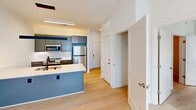 This well-designed one-bedroom, two-bathroom unit at 500 Maury offers a thoughtfully planned layout across a single floor. The open-concept kitchen serves as a central feature, showcasing grey cabinetry with sleek hardware, a bold blue island with a white quartz countertop, stainless steel appliances including a refrigerator, electric range, and built-in microwave, all set above light wood-tone flooring that runs throughout the unit. The living and entry areas flow naturally from the kitchen, creating a cohesive and functional common space bathed in clean, neutral tones. The bedroom is a bright, comfortable retreat featuring a ceiling fan, a window with natural light, and access to a dedicated walk-in closet with built-in shelving. Two bathrooms provide added convenience — one full bath features a backlit vanity mirror, grey cabinetry, white quartz countertop, and a soaking tub with shower, while the second bathroom mirrors the same modern aesthetic. A stacked in-unit washer and dryer is conveniently located near the secondary bathroom. Crisp white walls, light wood flooring, and consistent modern fixtures give the entire unit a clean, contemporary ambiance.