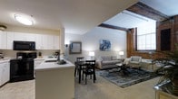 This single-floor space showcases a thoughtful blend of industrial character and contemporary design. The open-concept layout seamlessly connects the kitchen, dining, and living areas, creating a cohesive and functional living environment.

The kitchen features white cabinetry that extends to the ceiling, providing ample storage, and a light-colored countertop that doubles as a breakfast bar. The space is equipped with standard appliances, including a coffee maker, and the clean, neutral palette keeps the area feeling bright and organized.

The dining area sits just off the kitchen, accommodating a compact table with seating for four, making it ideal for casual meals or intimate gatherings. This area transitions naturally into the living room, which is anchored by a patterned area rug and furnished with a tufted sofa and accent chair.

One of the most striking architectural features is the exposed brick wall on one side of the living area, complemented by original wooden ceiling beams overhead. These industrial elements add warmth and texture, contrasting beautifully with the smooth white walls and modern furnishings. The vaulted ceiling height enhances the sense of openness throughout the space, while soft ambient lighting from floor and table lamps contributes to a welcoming atmosphere.