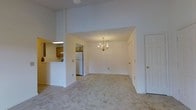 This single-floor residence offers a welcoming and open layout designed for comfortable everyday living. The main living and dining area is immediately impressive thanks to its soaring vaulted ceilings, which lend a generous sense of volume and airiness to the space. Soft, neutral-toned carpeting runs throughout, creating a warm and cohesive feel underfoot.

A elegant gold-toned chandelier serves as a focal point in the dining area, adding a classic decorative touch that complements the clean, white walls. Natural light filters in to brighten the space, enhancing its open and airy character.

The kitchen is positioned adjacent to the main living area and is accessible via a convenient pass-through counter, which encourages interaction between the kitchen and the living space while maintaining a degree of separation. The kitchen features warm wood-toned cabinetry and includes a full-size refrigerator, offering practical storage and functionality. An under-cabinet light provides task illumination over the work area.

Multiple doorways lead to additional rooms, suggesting a well-organized floor plan with clearly defined private and communal zones. Overall, the space presents a straightforward, functional layout with classic design details that provide a comfortable and inviting atmosphere throughout.