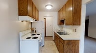 This single-floor space offers a practical and well-organized layout designed to maximize functionality. The galley-style kitchen is a standout feature, characterized by its warm honey-toned wood cabinetry that lines both sides of the corridor, providing generous storage space with upper and lower cabinet configurations. White appliances, including a coil-burner electric range and a full-size refrigerator, are neatly integrated along one wall, complemented by an overhead range hood. The opposite wall features a stainless steel sink set into light-colored countertops, with additional cabinetry above and below. The kitchen floor is finished with neutral tile, contributing to the clean and practical aesthetic of the space. Flush-mount ceiling lighting provides consistent illumination throughout. At the far end of the kitchen, a doorway leads to an adjoining area, suggesting a connected and accessible floor plan. The overall ambiance is functional and straightforward, with the warm wood tones of the cabinetry adding a cozy, traditional character to the space. This layout is well-suited for efficient meal preparation and everyday living.