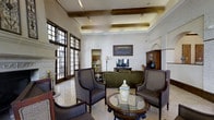 This single-story clubhouse presents a refined and welcoming interior characterized by classic architectural details and thoughtfully curated furnishings. The main lounge area showcases a coffered ceiling with exposed wooden beams and integrated ambient lighting, lending a sense of height and warmth to the space. Tall, multi-pane windows with dark wood frames line one wall, allowing generous natural light to filter in while adding a traditional architectural charm.

The seating area is arranged around a round glass-topped coffee table accented with a decorative ceramic centerpiece. Upholstered armchairs in neutral tones complement a deep olive-green sofa, creating an inviting conversational grouping. A dark wood entertainment unit anchors one wall, paired with a flat-screen television, while an arched alcove adds architectural interest nearby. A classic dark wood cabinet with glass-front doors provides additional storage and display space.

The light-toned tiled flooring ties the room together, reflecting the ambient light and enhancing the open feel. Wall art, a floor lamp, and subtle decorative accents complete the polished, traditional aesthetic, making this clubhouse a comfortable and elegant space for residents to gather and socialize.