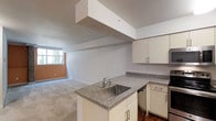 This single-floor space offers a thoughtfully designed open-concept layout that seamlessly connects the kitchen and living areas. The kitchen is outfitted with granite countertops featuring a speckled finish, a stainless steel sink set into a peninsula that doubles as a functional divider between the kitchen and main living space. White cabinetry with sleek brushed-metal hardware lines the kitchen walls, providing ample storage, while hardwood flooring grounds the culinary zone. A soffit detail above the peninsula adds architectural dimension to the ceiling, lending a structured, intentional design element to the space. The living area extends generously from the kitchen, finished with plush carpeting that adds warmth and comfort underfoot. A large window along the far wall invites an abundance of natural light, brightening the neutral-toned walls and creating an open, airy atmosphere throughout. The clean, minimalist aesthetic of the space—characterized by white walls, understated finishes, and a cohesive color palette—offers a versatile and move-in-ready environment suitable for a variety of furnishing styles and personal tastes.
