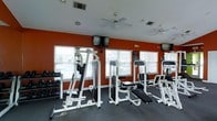 This fitness center offers a comprehensive workout environment designed to meet a range of exercise needs. The space is defined by its bold, deep-orange accent walls, which create an energizing atmosphere conducive to an active workout. The room is outfitted with multiple strength-training cable machines and resistance equipment, all finished in a clean white frame, providing a well-organized and visually cohesive layout. A dedicated free-weight section with a full dumbbell rack lines one side of the room, catering to users of all fitness levels. Overhead, dual ceiling fans help maintain a comfortable environment during intense workouts, while multiple windows fitted with horizontal blinds allow natural light to fill the space, giving it an open and airy feel. A wall-mounted flat-screen television is positioned prominently, allowing members to stay entertained during their sessions. The flooring features durable, dark carpeting suited for a gym environment. The single-floor layout ensures all amenities are easily accessible, making this fitness center a practical and well-appointed facility for daily exercise.