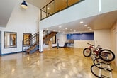 Located at 50 Drift Road in Breckenridge, Colorado, this two-story amenity space offers a well-designed and functional layout spread across two levels. The lower level serves as the primary activity floor, featuring polished concrete flooring throughout that lends a clean, industrial aesthetic. This level includes a dedicated bike storage and maintenance area, complete with wall-mounted bike racks, a repair stand, and tiled accent walls in white and blue that add a pop of color. A bright, open entry corridor with soaring double-height ceilings, glass-paneled exterior doors, and interior windows overlooking adjacent rooms connects the space and floods it with natural light. A striking open-tread staircase with steel cable railings and warm wood treads serves as both a functional and architectural centerpiece, rising to the upper mezzanine level. The mezzanine, finished with warm hardwood flooring, provides an open lounge or gathering area with industrial-style pendant lighting and steel cable railings that maintain visual connectivity to the floor below. Recessed lighting throughout both levels ensures consistent, even illumination. The overall design balances rugged mountain utility with modern, clean-lined finishes — well-suited to an active, outdoor-oriented community in the heart of the Colorado Rockies.