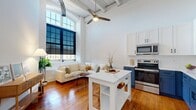 This thoughtfully designed one-bedroom, one-bathroom residence blends industrial character with modern finishes across a single floor. The open-concept living and kitchen area immediately impresses with dramatically high ceilings, exposed structural beams, and visible ductwork that nod to the building's architectural heritage. Rich, warm hardwood floors run throughout, anchoring the space with timeless appeal.

The kitchen is a standout feature, showcasing bold two-tone cabinetry with navy blue lower cabinets and crisp white uppers, complemented by white quartz countertops, a white subway tile backsplash, and a full suite of stainless steel appliances. An open wood shelf adds a casual, functional touch, while a freestanding island with seating provides additional prep and dining space.

The living area benefits from oversized industrial-style windows that invite generous natural light, framed by dark roller shades for privacy. A ceiling fan overhead adds to the comfort of the airy, loft-like atmosphere.

The bedroom is a serene, minimalist retreat with clean white walls, soaring ceilings, and continued hardwood flooring. The hallway features tall paneled doors with matte black hardware, adding a refined contrast to the bright surroundings.

The bathroom offers a sleek, modern aesthetic with large-format white wall tiles, bold black hexagonal mosaic floor tiles, a glass-enclosed walk-in shower, and a clean white vanity.