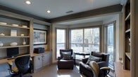 This well-appointed single-floor space exudes a refined, intimate atmosphere through its thoughtful design and carefully chosen details. The sitting area serves as the focal point, anchored by a pair of plush dark brown leather armchairs accented with teal throw pillows, arranged to encourage comfortable conversation or quiet relaxation. A built-in cabinetry unit lines one wall, featuring a wall-mounted display screen, integrated shelving with decorative fluted column details, and lower cabinet storage — a combination that is both functional and visually cohesive. The neutral color palette of warm grays and taupe tones, complemented by dark crown molding and ceiling beams, lends the space a sophisticated, classic character. A bank of large bay windows floods the room with generous natural light, framing views of mature trees and the surrounding outdoor landscape, creating a seamless connection between the interior and nature. Recessed ceiling lighting provides warm, even illumination throughout. The textured neutral carpeting underfoot adds a layer of comfort and warmth, tying together the room's elegant yet welcoming aesthetic. Overall, this space balances practicality with style, making it ideal for both focused work and leisurely relaxation.