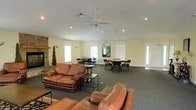 This single-floor communal space is thoughtfully designed to accommodate both relaxed lounging and social gatherings. The open-concept layout is anchored by a striking stone fireplace on one wall, adding warmth and a rustic focal point to the room. Rich brown leather sofas and loveseats are arranged around the fireplace area, creating an inviting conversation and relaxation zone, complemented by low-profile coffee and side tables.

The space transitions seamlessly into a dining and game area, where multiple round tables with dark wooden chairs offer ample seating for group activities or casual meals. Natural light filters in through several windows fitted with white blinds, keeping the atmosphere bright and open. A decorative arched mirror adds an elegant touch to the wall décor.

Overhead, a ceiling fan with integrated lighting ensures comfortable air circulation throughout the room, supported by recessed lighting for well-distributed illumination. The dark, low-pile carpeted flooring ties the space together cohesively. A clearly marked emergency exit door and an entry door with sidelight window reinforce the functional, safety-conscious design of this well-appointed shared space.