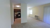 Located at 8670 Braewood Dr in Baldwinsville, New York, this single-floor residence offers a practical and comfortable layout designed for easy everyday living. The living area features neutral wall-to-wall carpeting, soft yellow-toned walls, and flush-mount ceiling lighting that creates a warm, evenly lit environment. A wall-mounted air conditioning unit and a baseboard heater ensure year-round climate control, while a window allows for natural light to enter the space. The kitchen is separated from the living area by a partial wall, providing a sense of division while maintaining an open feel. It is outfitted with dark wood cabinetry, black appliances including a gas range and refrigerator, and a practical layout suited for home cooking. The overall design is straightforward and functional, with clean lines and a neutral color palette throughout. The single-level floor plan offers accessibility and ease of movement between spaces, making this residence well-suited for a variety of occupants seeking a manageable, low-maintenance home in the Baldwinsville area of Central New York.