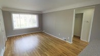 This single-floor space presents a straightforward and functional layout, characterized by its clean, minimalist aesthetic. The main living area is an open, generously proportioned room finished with warm-toned hardwood flooring that runs throughout, adding a natural warmth to the interior. The walls are painted in a neutral grey tone, complemented by crisp white baseboards and trim that provide a clean contrast.

A large window fitted with vertical blinds allows natural light to filter into the space, illuminating the room and offering views of the surrounding greenery outside. The window's placement enhances the bright, airy feel of the interior. A floor-mounted vent indicates a central heating and cooling system for year-round climate control.

A doorway is visible along one wall, hinting at additional rooms or storage space beyond the main living area. The overall layout is unobstructed and versatile, providing flexibility for various furniture arrangements. The combination of neutral tones, natural wood flooring, and ample natural light gives the space a calm, welcoming ambiance suited to a variety of uses.