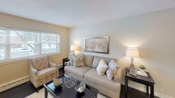 This single-floor unit offers a straightforward and functional layout, as revealed by the 3D floor plan, which shows distinct living, sleeping, and kitchen areas connected by an efficient corridor. The living area is generously proportioned, featuring rich, dark hardwood flooring that runs throughout the main rooms. Neutral beige walls and white trim provide a clean, understated backdrop, while baseboard heating units line the perimeter, ensuring consistent warmth. Windows allow natural light to filter into the space, offering views of surrounding greenery.

The bedroom is finished with wall-to-wall carpet in a warm taupe tone, contributing a softer feel underfoot. A ceiling fan with a built-in light fixture provides both overhead lighting and air circulation. A single window with blinds brings in natural light while maintaining privacy, and baseboard heating continues here as well.

The kitchen follows a galley-style configuration, maximizing efficiency within a compact footprint. Gray raised-panel cabinetry pairs with granite-look countertops and a white subway tile backsplash. White appliances, including an electric range and refrigerator, are included, and a window above the sink area brightens the workspace. Tile flooring completes the kitchen's practical, easy-to-maintain design.