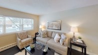 This single-floor unit offers a straightforward and functional layout, as revealed by the 3D floor plan, which shows distinct living, sleeping, and kitchen areas connected by an efficient corridor. The living area is generously proportioned, featuring rich, dark hardwood flooring that runs throughout the main rooms. Neutral beige walls and white trim provide a clean, understated backdrop, while baseboard heating units line the perimeter, ensuring consistent warmth. Windows allow natural light to filter into the space, offering views of surrounding greenery.

The bedroom is finished with wall-to-wall carpet in a warm taupe tone, contributing a softer feel underfoot. A ceiling fan with a built-in light fixture provides both overhead lighting and air circulation. A single window with blinds brings in natural light while maintaining privacy, and baseboard heating continues here as well.

The kitchen follows a galley-style configuration, maximizing efficiency within a compact footprint. Gray raised-panel cabinetry pairs with granite-look countertops and a white subway tile backsplash. White appliances, including an electric range and refrigerator, are included, and a window above the sink area brightens the workspace. Tile flooring completes the kitchen's practical, easy-to-maintain design.