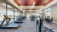 Located at 12479 Rojas Dr in Horizon City, TX, this well-appointed fitness center offers a comprehensive workout facility across a single open floor. The space is defined by its striking architectural design, highlighted by coffered ceilings with bold terracotta-red insets, multiple ceiling fans, and recessed and linear lighting that ensure the space is bright and inviting throughout the day. A defining feature is the expansive floor-to-ceiling window wall on one side, flooding the interior with natural light and providing open views of the surrounding community. The flooring throughout is durable, commercial-grade carpet, providing comfort underfoot across all workout zones. Cardio equipment—including treadmills, ellipticals, and a rowing machine—is positioned along the windowed wall to take advantage of the outdoor views. The strength training area is well-equipped with cable machines, weight racks, benches, and multi-function resistance stations. A mounted flat-screen television keeps members engaged during workouts. A vibrant geometric accent wall in warm tones of red, gold, and cream adds a dynamic visual focal point and reinforces the modern aesthetic of the space. Clerestory windows along additional walls further contribute to the airy, open atmosphere of this thoughtfully designed fitness facility.
