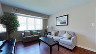 This single-floor residence offers a thoughtfully arranged layout with consistent warm-toned walls and rich hardwood flooring across the main living areas. The living room is a welcoming, well-lit space featuring large windows with vertical blinds that allow generous natural light to fill the room. It comfortably accommodates a full sofa and loveseat arrangement centered around a dark wood coffee table, with a flat-screen TV and wall art completing the space.

Adjacent to the living room, the dining area is defined by a pendant light overhead and classic chair rail molding along the walls. A dark wood dining table with four matching upholstered chairs provides an intimate setting for meals. Framed artwork and a decorative plant accent the corner.

The kitchen is compact yet highly functional, featuring rich espresso-toned cabinetry, granite-style countertops, and a full suite of stainless steel appliances including a range, microwave, and dishwasher. A window above the sink provides natural light.

The bedroom is a serene retreat with plush carpeting, dual windows offering ample daylight, and sufficient space for a queen bed and nightstand. Framed wall art and a tray ceiling detail add a refined touch.

The bathroom maintains a clean, neutral aesthetic with light tile flooring, a white vanity, and a simple, functional layout that completes this well-rounded residence.