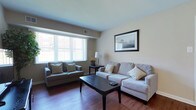 This single-floor residence offers a thoughtfully arranged layout with consistent warm-toned walls and rich hardwood flooring across the main living areas. The living room is a welcoming, well-lit space featuring large windows with vertical blinds that allow generous natural light to fill the room. It comfortably accommodates a full sofa and loveseat arrangement centered around a dark wood coffee table, with a flat-screen TV and wall art completing the space.

Adjacent to the living room, the dining area is defined by a pendant light overhead and classic chair rail molding along the walls. A dark wood dining table with four matching upholstered chairs provides an intimate setting for meals. Framed artwork and a decorative plant accent the corner.

The kitchen is compact yet highly functional, featuring rich espresso-toned cabinetry, granite-style countertops, and a full suite of stainless steel appliances including a range, microwave, and dishwasher. A window above the sink provides natural light.

The bedroom is a serene retreat with plush carpeting, dual windows offering ample daylight, and sufficient space for a queen bed and nightstand. Framed wall art and a tray ceiling detail add a refined touch.

The bathroom maintains a clean, neutral aesthetic with light tile flooring, a white vanity, and a simple, functional layout that completes this well-rounded residence.