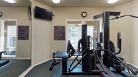 This fitness center is a thoughtfully organized exercise facility spread across a single floor, designed to accommodate a range of workout needs. The space is divided into interconnected areas, allowing for a natural flow between cardio and strength training zones.

The main exercise area features dark carpet flooring throughout, providing a comfortable and functional surface underfoot. A large wall-mounted mirror spans a significant portion of one wall, visually expanding the room and allowing users to monitor their form during workouts. Flush-mounted ceiling fixtures provide consistent, even lighting across the space.

Strength training is supported by a Vectra multi-station cable machine, complemented by a weight bench, making the area suitable for a variety of resistance exercises. Cardio equipment includes a treadmill, an elliptical trainer, and a recumbent stationary bike, offering multiple options for aerobic exercise. A wall-mounted flat-screen television is positioned for easy viewing during workouts.

Natural light enters through windows and a glass door that leads to an outdoor area, adding brightness to the space. A wall clock and a posted fitness center rules board complete the functional, community-oriented character of the facility.
