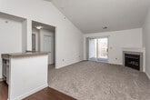 This single-floor residence is characterized by its vaulted ceilings in the main living areas, which lend a sense of openness and volume to the space. The living room is a standout feature, showcasing soaring angled ceilings, a ceiling fan, neutral carpet flooring, a tiled fireplace, and sliding glass doors that open to a private balcony with outdoor views. The open layout connects the living area to the kitchen, which is defined by a peninsula counter with a laminate surface, white cabinetry, and dark hardwood-style flooring. The kitchen is well-equipped with a range, built-in microwave, refrigerator, and dishwasher, with natural light filtering in from a window at the end of the galley-style corridor. The bedroom features its own ceiling fan, a large window that brings in natural light, and plush neutral carpet. The bathroom offers a single-sink vanity with laminate countertop and wood-toned cabinetry, a large mirror with overhead lighting, a combination tub and shower, and tile flooring. Throughout the home, white walls and neutral finishes create a clean, consistent aesthetic suited for a variety of décor styles.
