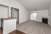 This single-floor residence is characterized by its vaulted ceilings in the main living areas, which lend a sense of openness and volume to the space. The living room is a standout feature, showcasing soaring angled ceilings, a ceiling fan, neutral carpet flooring, a tiled fireplace, and sliding glass doors that open to a private balcony with outdoor views. The open layout connects the living area to the kitchen, which is defined by a peninsula counter with a laminate surface, white cabinetry, and dark hardwood-style flooring. The kitchen is well-equipped with a range, built-in microwave, refrigerator, and dishwasher, with natural light filtering in from a window at the end of the galley-style corridor. The bedroom features its own ceiling fan, a large window that brings in natural light, and plush neutral carpet. The bathroom offers a single-sink vanity with laminate countertop and wood-toned cabinetry, a large mirror with overhead lighting, a combination tub and shower, and tile flooring. Throughout the home, white walls and neutral finishes create a clean, consistent aesthetic suited for a variety of décor styles.