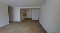 This single-floor space presents an open and functional layout with a straightforward design. The main living area is finished with wall-to-wall carpeting in a neutral beige tone, creating a cohesive and understated aesthetic. White walls throughout contribute to a clean, bright atmosphere, while a ceiling-mounted light fixture provides overhead illumination.

A structural beam or soffit defines the transition between the living area and the adjoining kitchen space, which is partially visible from the main room. The kitchen features darker countertops that contrast with the lighter cabinetry and walls, and overhead lighting keeps the area well-lit. A passthrough or open wall connects the kitchen to the living space, promoting an open feel between the two areas.

To the right of the main living area, a hallway leads further into the unit, suggesting access to additional rooms. A ceiling vent is visible, indicating a central HVAC system. The overall design is practical and minimal, with neutral finishes that offer a versatile backdrop suitable for a variety of furnishing styles.