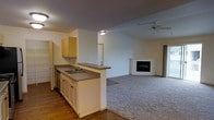 This single-story space is designed around an open-concept layout that connects the kitchen and living area into one cohesive environment. The kitchen is positioned along a peninsula counter with a double-basin sink, white lower cabinetry with dark hardware, and light wood upper cabinets mounted above. The countertop extends outward, creating a natural divider between the kitchen and the adjoining living space while still maintaining visual openness throughout.

The flooring transitions from dark hardwood in the kitchen zone to plush carpeting in the living area, clearly delineating the two functional spaces. The living area is generously sized, anchored by a corner fireplace with a clean, minimalist surround that serves as a focal point. A ceiling fan is mounted overhead, providing both airflow and a practical lighting fixture for the room.

The vaulted ceiling adds a sense of height and openness to the space, enhancing the airy feel of the interior. Neutral wall tones and minimal detailing give the space a clean, versatile aesthetic. A sliding glass door on one side of the living area suggests access to an outdoor area, allowing for natural light to filter in.