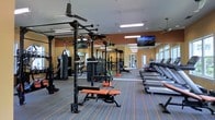 This single-floor fitness center offers a comprehensive workout environment designed to accommodate a wide range of exercise routines. The space is organized into two primary zones: a strength training area and a dedicated cardio section. The strength zone features a multi-functional power rack system complete with cable attachments, a heavy bag for boxing training, a weight bench with orange upholstery, and various resistance and functional fitness equipment. The cardio section lines the perimeter with a row of treadmills positioned near large windows, allowing natural light to fill the space during workouts. Mirrored walls run along one side of the room, enhancing the sense of openness and allowing users to monitor their form. The flooring consists of durable, patterned carpet with orange accent stripes that complement the warm orange tones of the walls. Overhead, ceiling fans and fluorescent lighting fixtures ensure the space remains well-ventilated and brightly lit. Wall-mounted flat-screen televisions are positioned for easy viewing from both the strength and cardio areas. The overall ambiance is energetic and functional, reflecting a thoughtfully designed space built to support a full range of fitness activities.