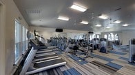 This fitness center offers a comprehensive workout environment designed to accommodate a variety of exercise routines. The space is generously sized, providing ample room for members to move freely between equipment without feeling crowded. Rows of cardio machines, including treadmills, ellipticals, and stationary bikes, are arranged along one side of the room, while strength-training machines occupy the remaining floor space. A large wall-to-wall mirror runs along one side, enhancing the sense of openness and allowing users to monitor their form during workouts. The flooring features a striped, multi-toned carpet that adds a modern touch while providing a degree of cushioning underfoot. Overhead, multiple ceiling-mounted fans work alongside bright panel lighting to maintain a comfortable and well-lit environment throughout the space. The neutral gray walls and clean layout contribute to a focused, functional atmosphere. Natural light filters in through windows along the perimeter, further brightening the interior. The overall design prioritizes practicality and accessibility, making this fitness center a well-rounded facility suited for both cardio-focused and strength-training workouts.