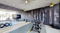 This dynamic fitness facility is designed to energize and inspire, offering a well-equipped workout environment across a single floor. The space makes an immediate visual impact with a bold, large-scale black-and-white photographic mural of the Charlotte city skyline, serving as a dramatic backdrop that sets a motivating tone throughout the area. A row of professional-grade spinning bikes is positioned front and center, ideal for high-intensity cycling sessions. A large flat-screen display mounted on the mirrored wall provides access to Fitness on Demand, enabling members to follow guided workout classes at any time. The full-length mirrors along the wall not only amplify the sense of space but also allow users to monitor their form during training. Overhead, a ceiling fan ensures comfortable air circulation during intense workouts, complemented by recessed lighting and pendant fixtures that provide a bright, well-lit environment. The flooring features a combination of striped carpet and functional fitness surfaces, contributing to both the aesthetic appeal and practical utility of the space. The overall ambiance is modern and urban, combining high-end finishes with purposeful fitness infrastructure to deliver a premium workout experience.