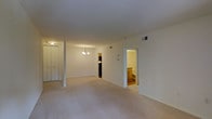 This single-floor residence offers a clean, functional layout designed for comfortable everyday living. The main living area is generously proportioned, featuring soft wall-to-wall carpeting in a neutral tone that lends warmth and continuity throughout the space. Crisp white walls and a smooth ceiling create a bright, airy atmosphere, while HVAC vents are thoughtfully integrated into both the walls and ceiling to ensure year-round comfort. A structural column adds subtle architectural definition to the open floor plan without interrupting the sense of spaciousness. A classic chandelier serves as a focal point, casting warm ambient light across the room and adding a touch of elegance to the otherwise understated interior. Built-in closet storage with full-height bifold doors lines one wall, providing ample organization space. Adjacent to the main living area, a doorway leads into a well-appointed bathroom finished with warm wood cabinetry and traditional fixtures, offering a functional and private retreat. A secondary doorway hints at additional rooms or storage areas beyond, suggesting a thoughtfully connected interior layout. Overall, the space presents a versatile, move-in-ready environment with a neutral palette suited to a variety of furnishing styles.