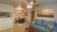 This single-floor residence offers a welcoming and functional layout centered around an open-concept living and dining area. The living space is anchored by a comfortable sofa in a soft blue tone, paired with a wooden coffee table and accented by decorative wall art that adds warmth and character to the room. A five-blade ceiling fan with an integrated light fixture provides both comfort and ambient lighting overhead.

Adjacent to the living area, the dining space features a classic round table surrounded by dark wood chairs, set atop a plush area rug. A chandelier hangs above, adding a touch of elegance to the dining experience. The seamless flow between the two areas creates a cohesive and inviting atmosphere ideal for both everyday living and entertaining.

Throughout the space, neutral-toned walls and carpeted flooring contribute to a cozy, understated aesthetic. A doorway visible from the main living area hints at additional private rooms beyond, suggesting a practical and well-organized floor plan. The overall ambiance is warm, comfortable, and thoughtfully arranged, making it an approachable and livable space.