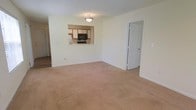 This single-floor unit features an open and functional layout highlighted by a spacious carpeted living area finished in neutral beige tones. The soft carpeting extends throughout the main living space, contributing to a warm and comfortable atmosphere. A pass-through window connects the living area to the kitchen, creating an open sightline that enhances the sense of space. The kitchen is equipped with oak-toned cabinetry, a microwave, and black appliances, offering a practical and efficient cooking space. White walls and white trim throughout the unit provide a clean, bright aesthetic, complemented by flush-mounted ceiling lighting that ensures even illumination. A white six-panel door leads to an additional room, suggesting a private and well-defined layout. A doorway visible along the left wall provides access to additional areas of the unit. The overall design is straightforward and practical, with a neutral color palette that offers flexibility for a variety of furnishing styles.